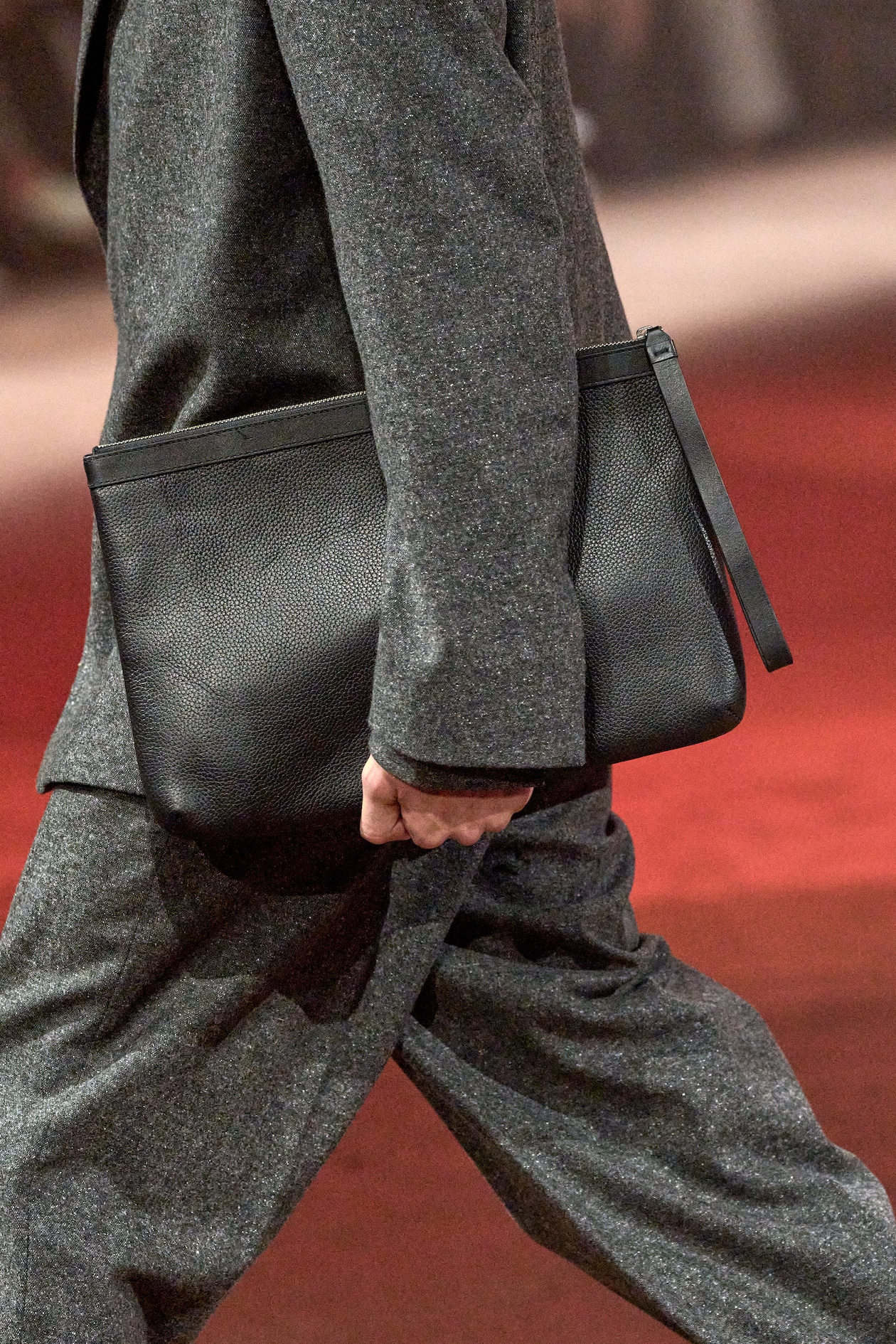 Model wears grey coat, grey pants and holds black leather bag