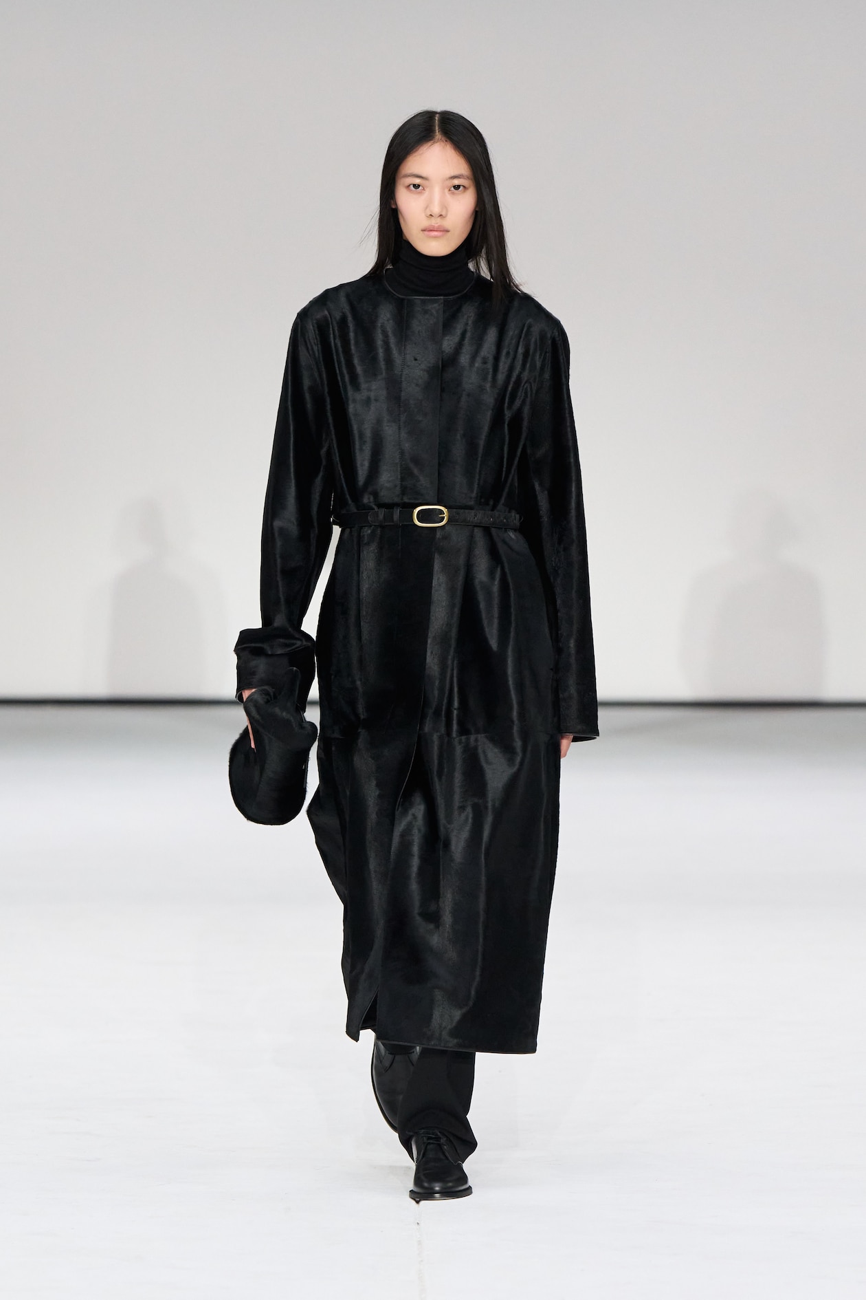 Model in a long black coat with a belt, black turtleneck, and holding a black hat, walks on a runway against a plain background.