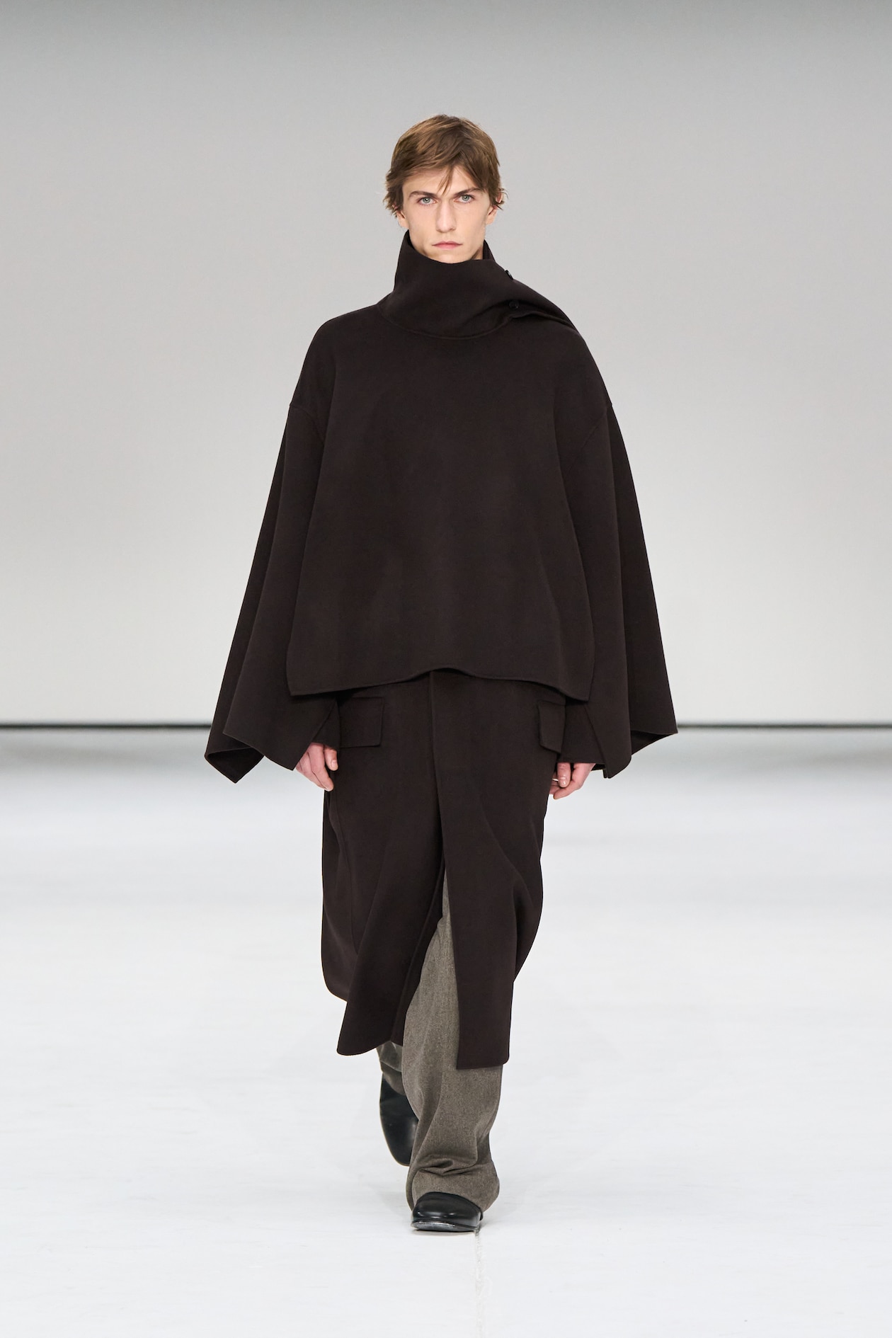 Person wearing a dark, oversized poncho-style outfit with wide pants and black shoes, walking on a minimalist white runway.