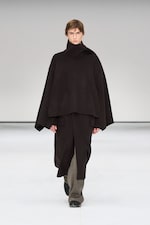 Placeholder for: Person wearing a dark, oversized poncho-style outfit with wide pants and black shoes, walking on a minimalist white runway.