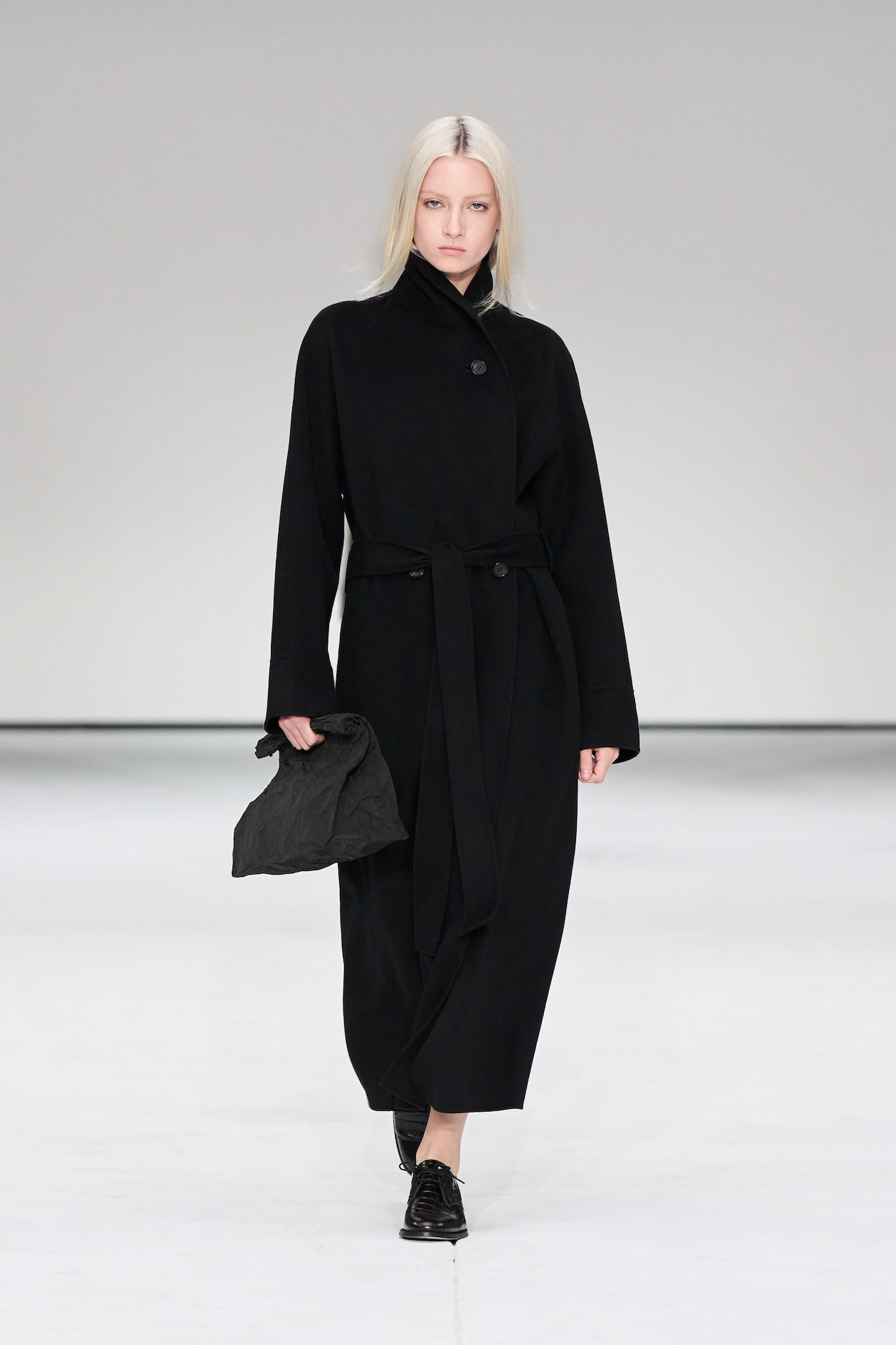 Model in a long black coat and black shoes walks on a white runway, holding a dark clutch, against a neutral background.