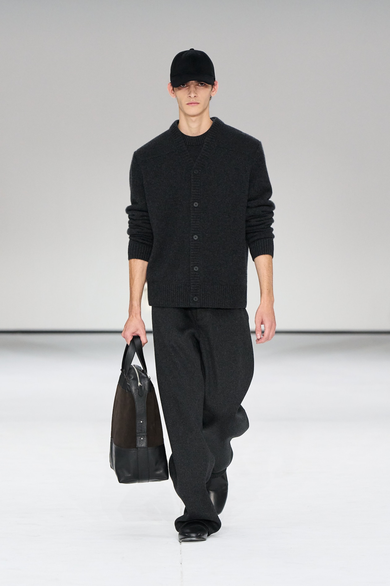 Model in a dark outfit with a black cap and bag walks on a white runway.