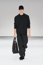 Placeholder for: Model in a dark outfit with a black cap and bag walks on a white runway.