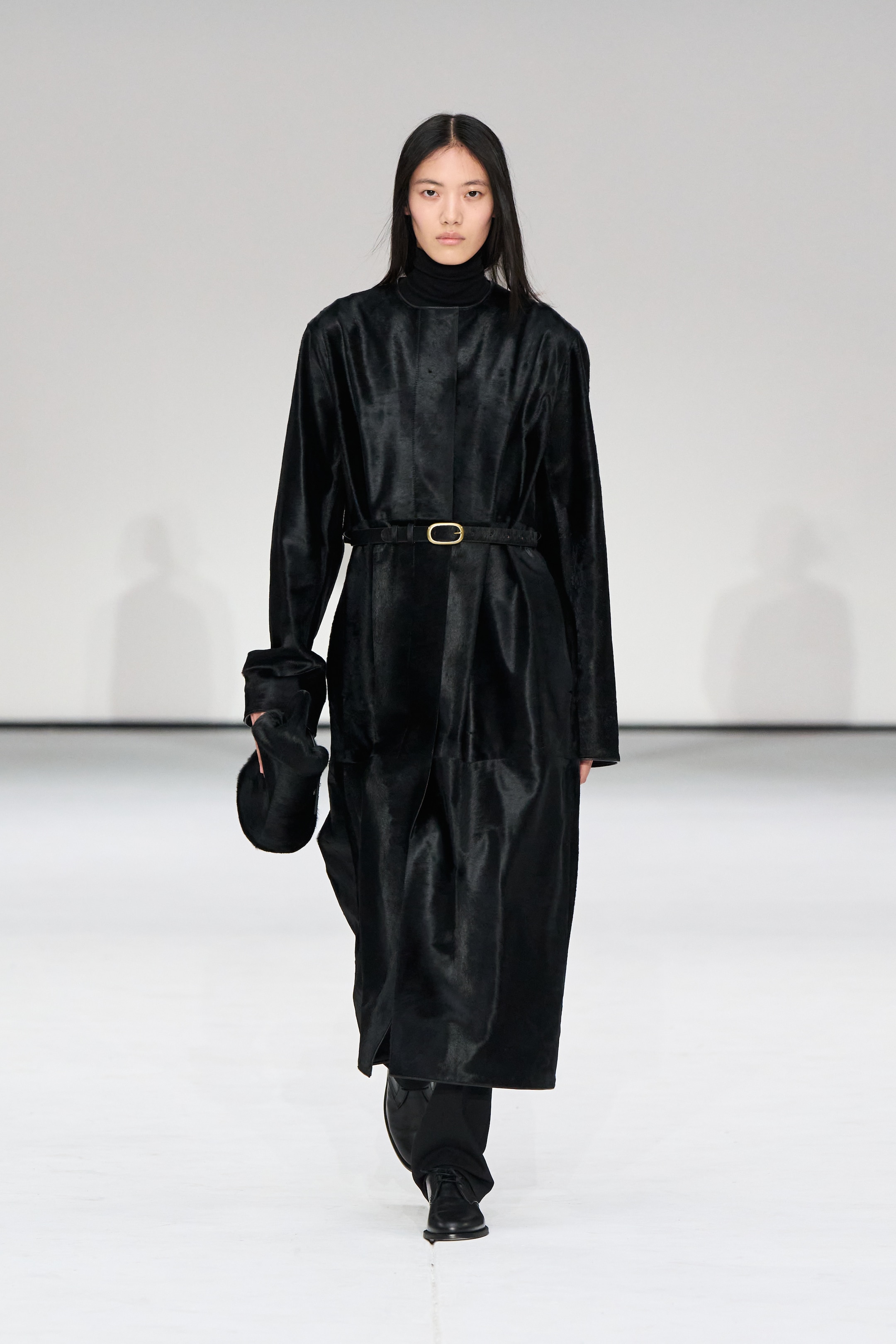 Model in a long black coat with a belt, black pants, and boots, holding a black hat, walks on a white runway.