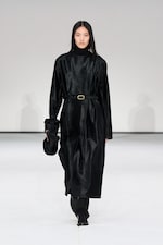 Placeholder for: Model in a long black coat with a belt, black pants, and boots, holding a black hat, walks on a white runway.
