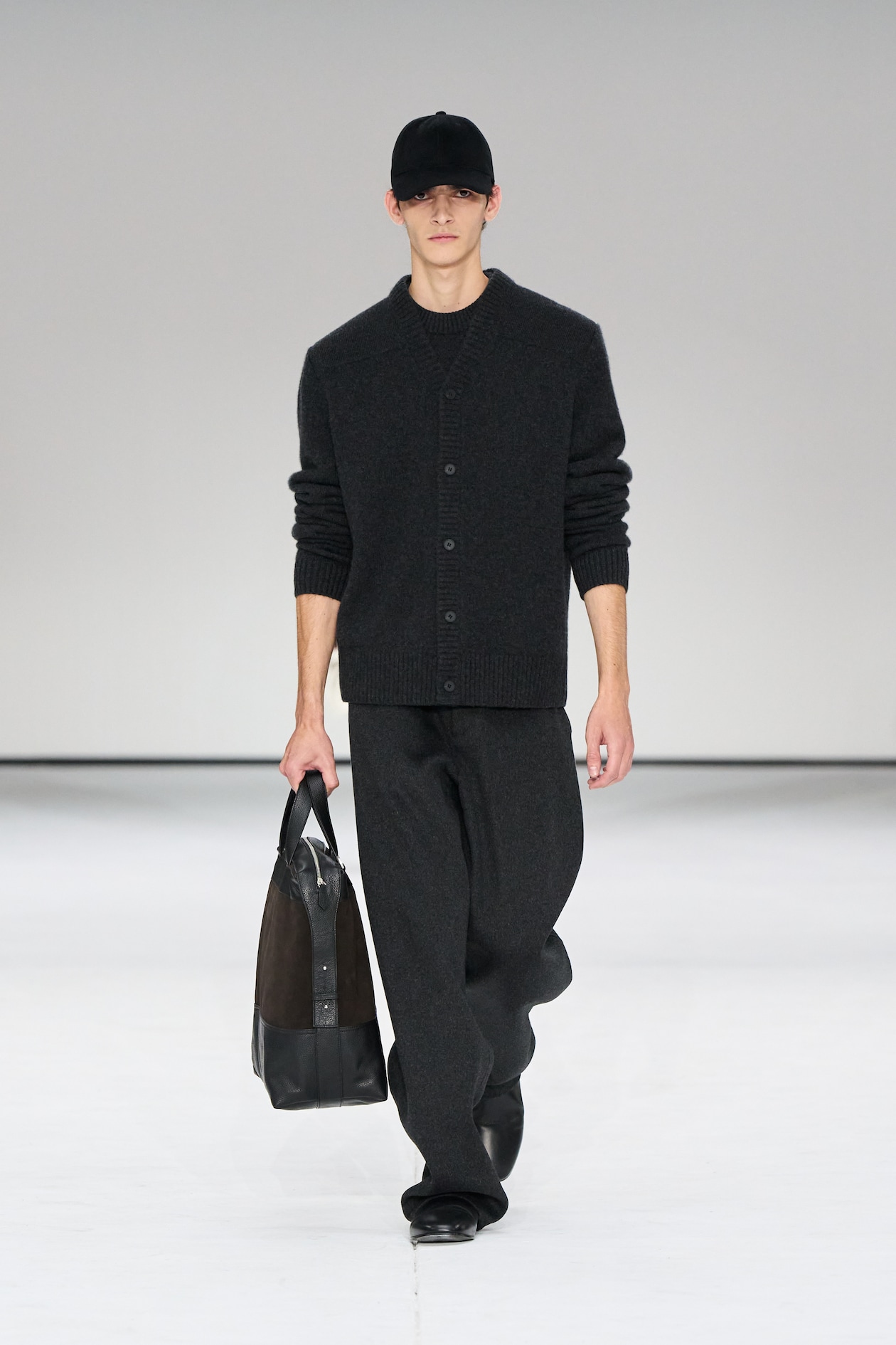 Person in a dark outfit, including a cap and sweater, walks on a runway holding a black bag. White background.