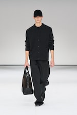 Placeholder for: Person in a dark outfit, including a cap and sweater, walks on a runway holding a black bag. White background.