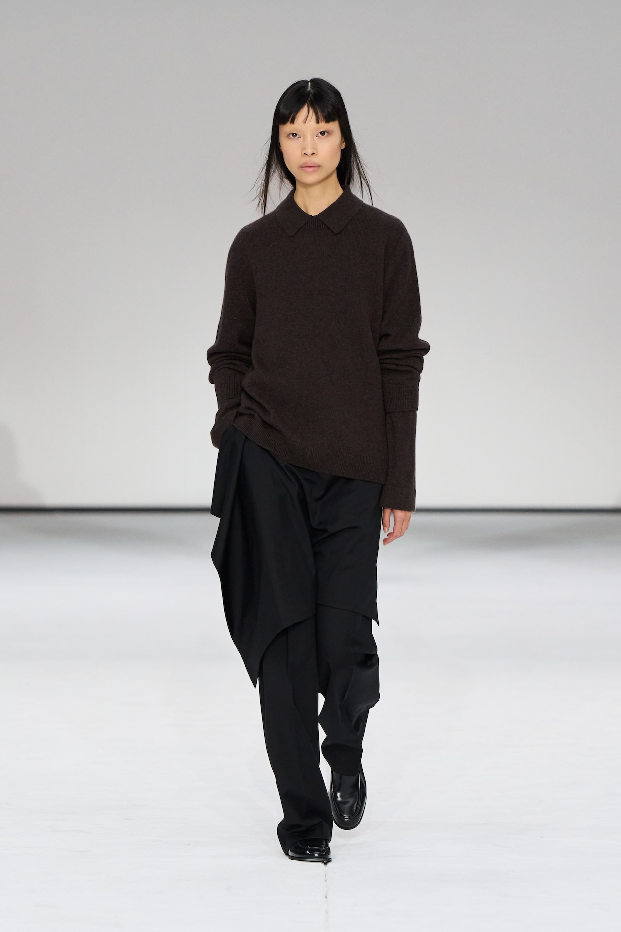 Model in a brown sweater and black pants walks on a runway against a plain white background.