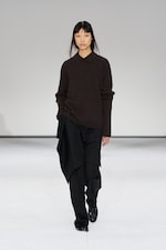 Placeholder for: Model in a brown sweater and black pants walks on a runway against a plain white background.