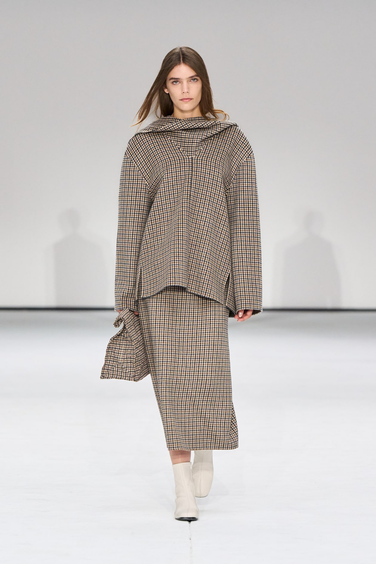 Model in a plaid outfit walks on a white runway, wearing a matching top and skirt with white ankle boots.