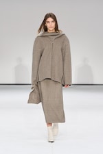 Placeholder for: Model in a plaid outfit walks on a white runway, wearing a matching top and skirt with white ankle boots.