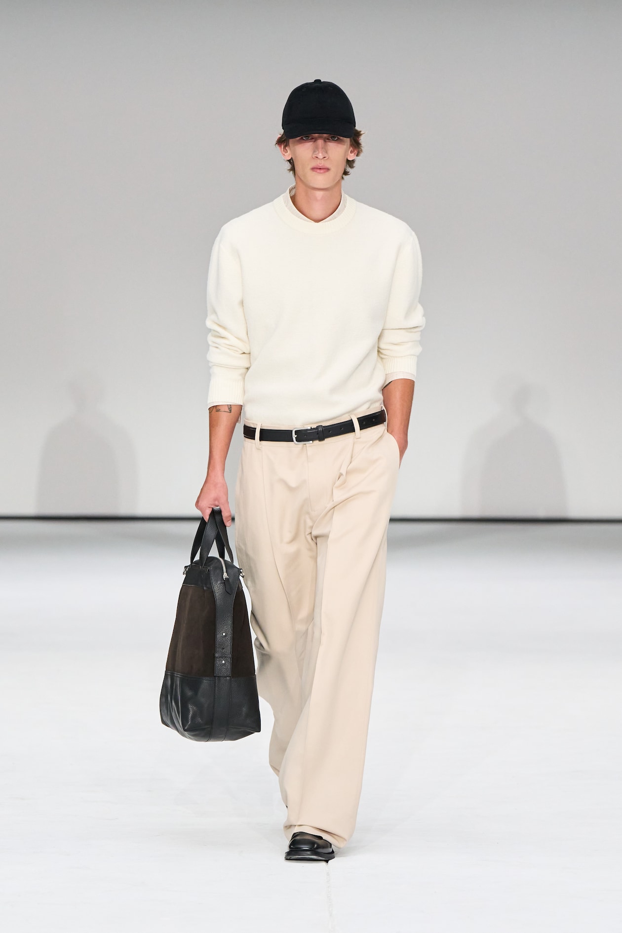 Model in a white sweater, beige pants, and black cap walks on runway carrying a black leather bag.