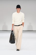 Placeholder for: Model in a white sweater, beige pants, and black cap walks on runway carrying a black leather bag.