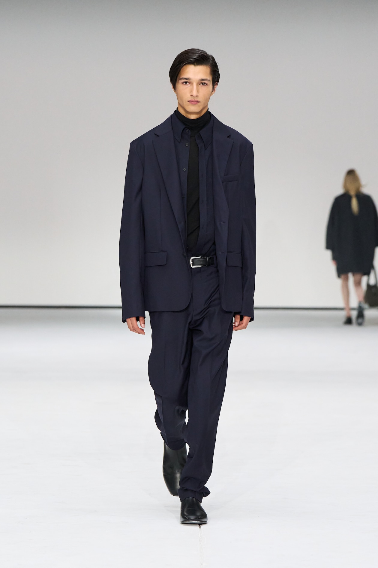 Model in a dark blue oversized suit and black shirt walks on a white runway, with another person blurred in the background.
