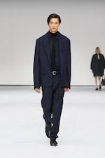 Placeholder for: Model in a dark blue oversized suit and black shirt walks on a white runway, with another person blurred in the background.