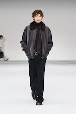 Placeholder for: Model in a black leather jacket and trousers walks on a white runway, hands in pockets, with a blurred figure in the background.