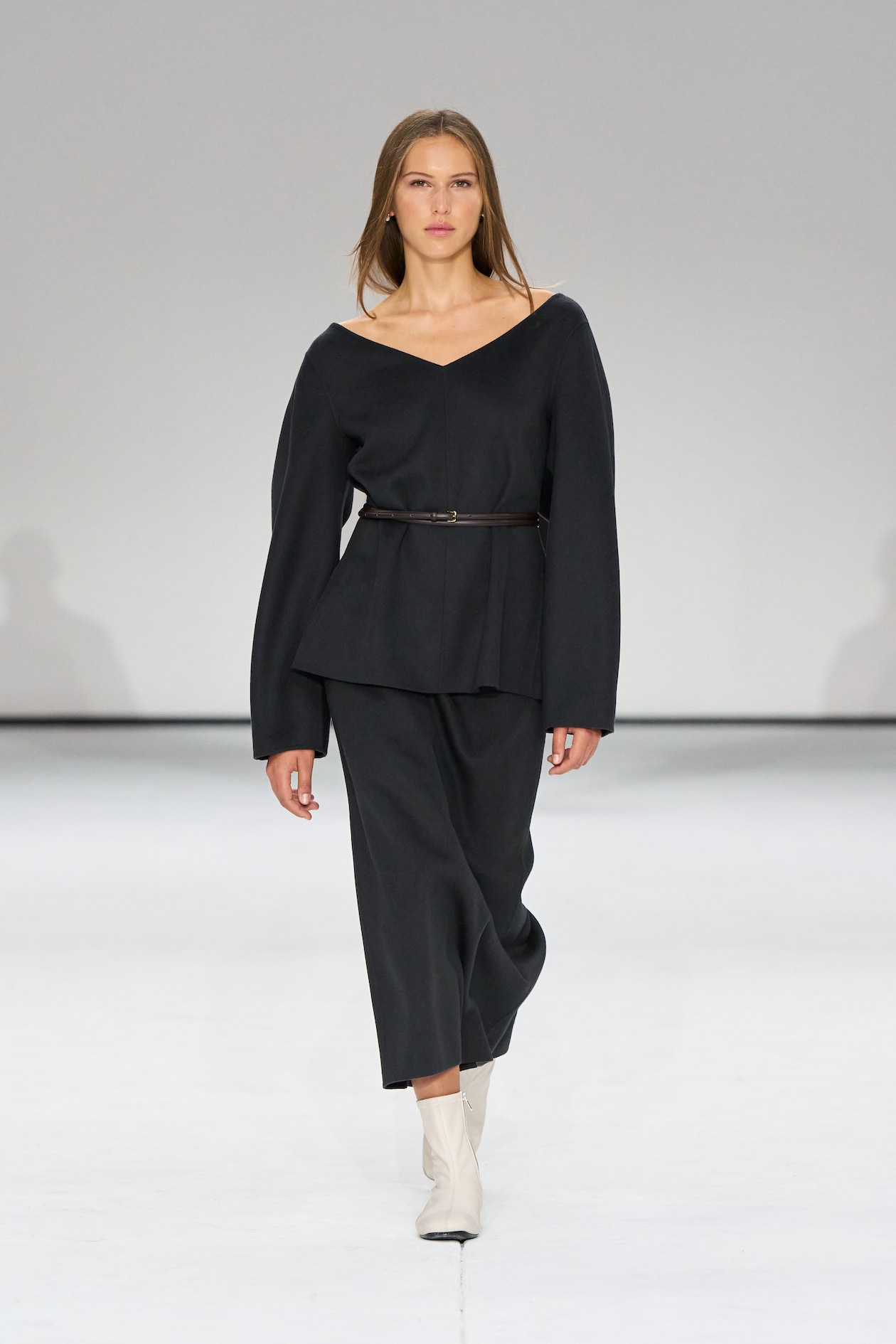 Model walking on a runway wearing a dark, off-shoulder outfit with a belt, paired with white ankle boots. Neutral background.