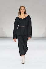 Placeholder for: Model walking on a runway wearing a dark, off-shoulder outfit with a belt, paired with white ankle boots. Neutral background.