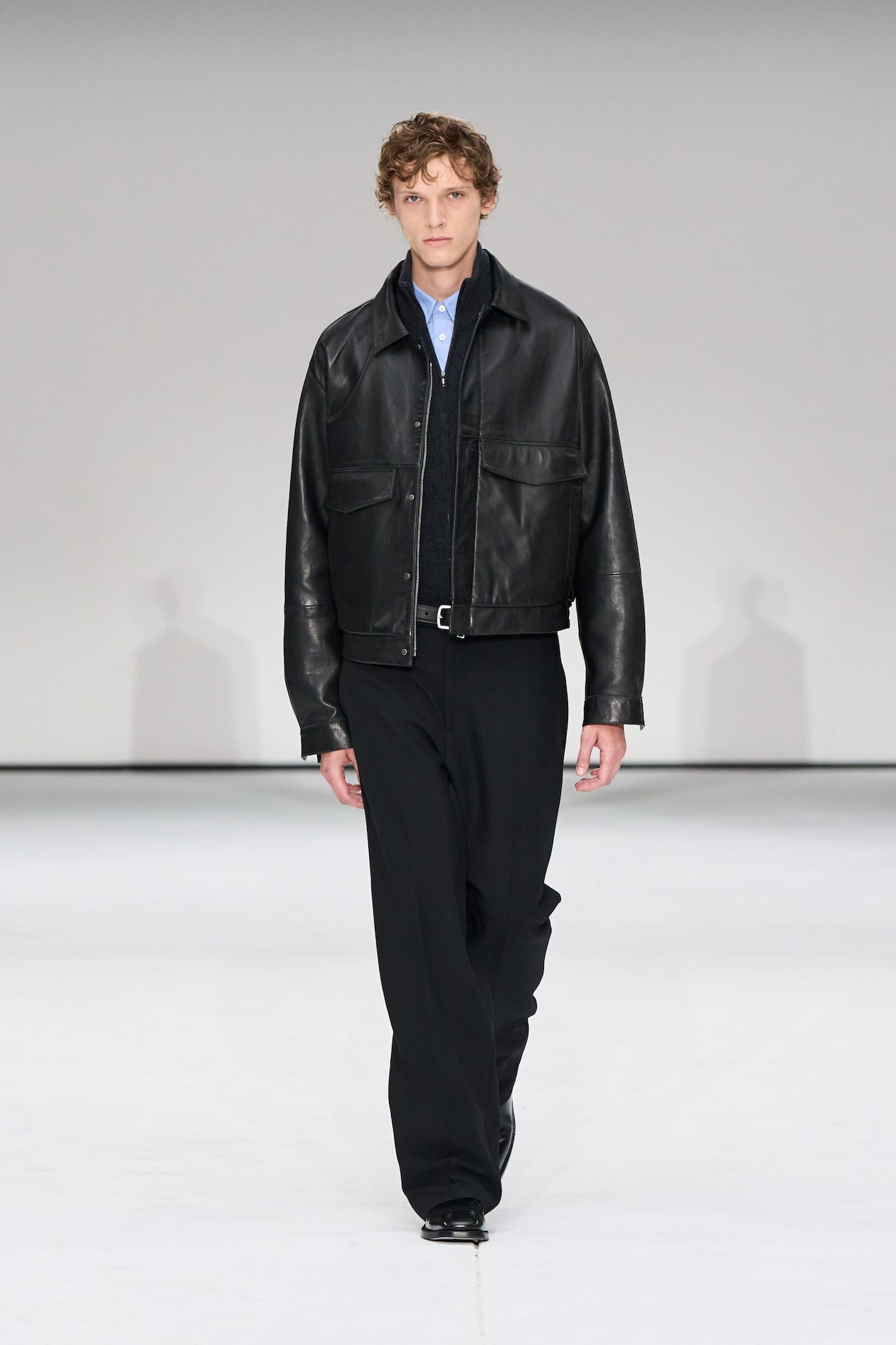 A person walks on a runway wearing a black leather jacket, blue shirt, and black pants against a plain background.