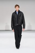 Placeholder for: A person walks on a runway wearing a black leather jacket, blue shirt, and black pants against a plain background.