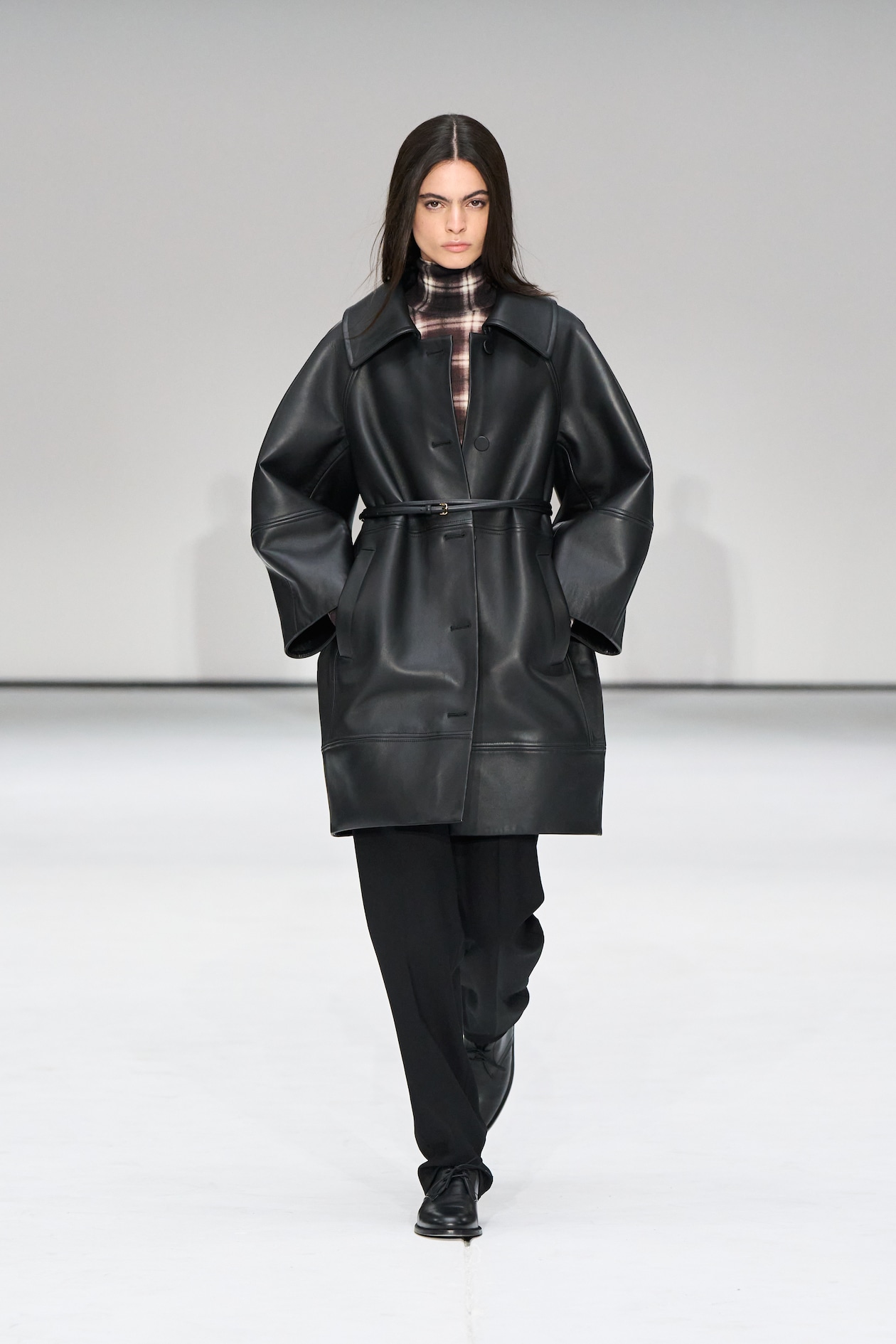 Model in a black leather coat with a plaid top and black pants walks on a runway against a plain background.