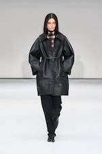 Placeholder for: Model in a black leather coat with a plaid top and black pants walks on a runway against a plain background.