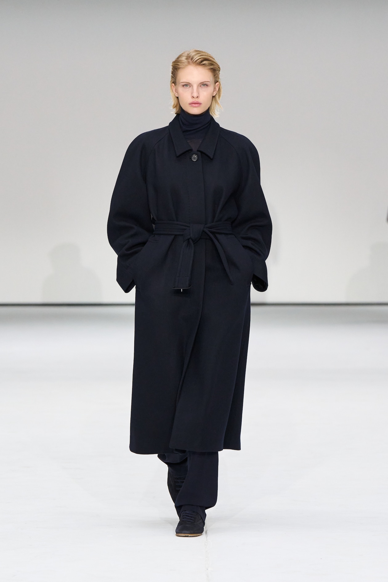 Person walking on a runway wearing a long, dark coat with a belt, black pants, and a turtleneck, against a plain background.