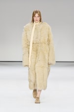 Placeholder for: Model wearing a long, fluffy beige fur coat with a matching scarf and brown shoes, walking on a white runway.