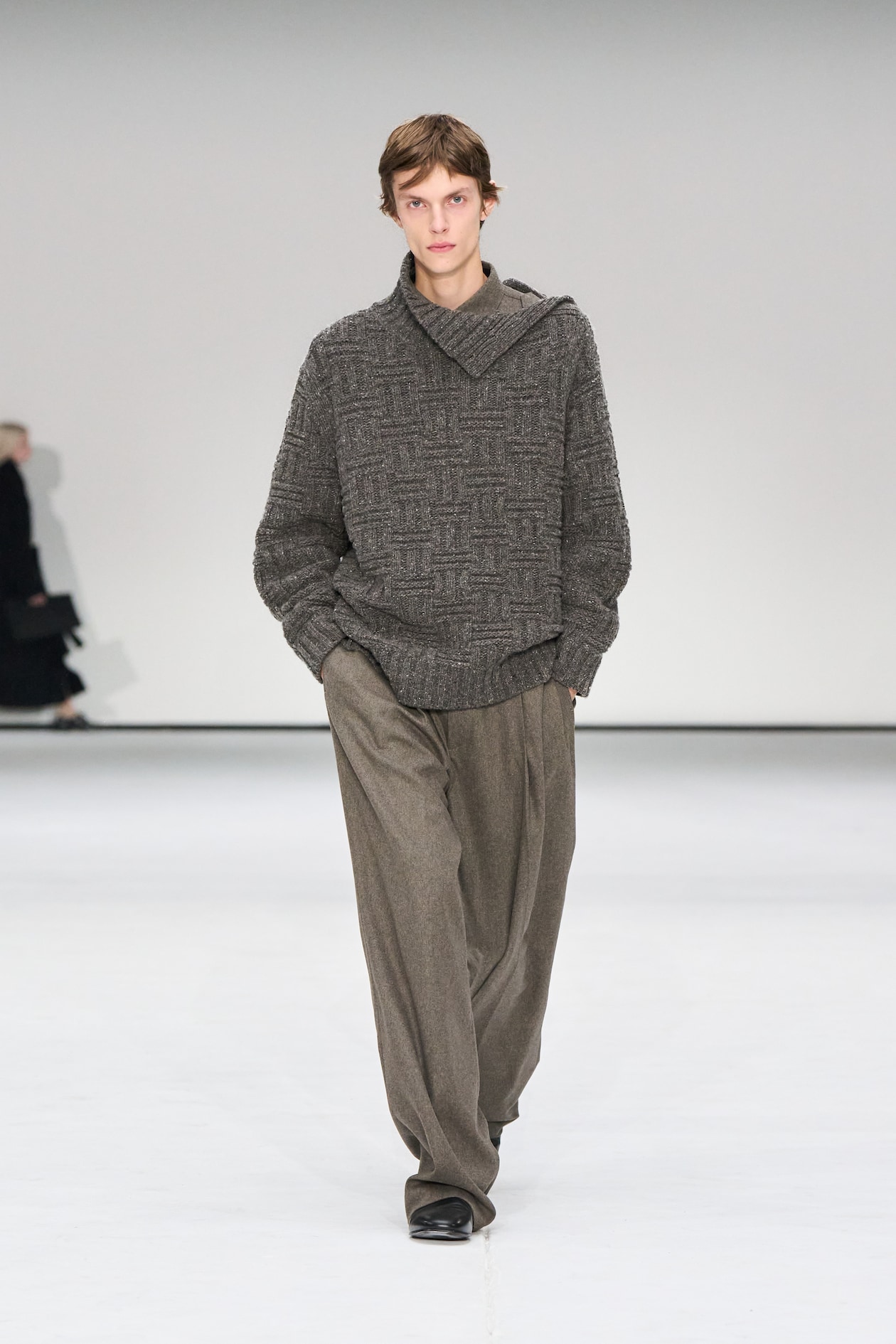 Person walking on a runway wearing a textured gray sweater and loose-fitting gray trousers, with a plain white background.