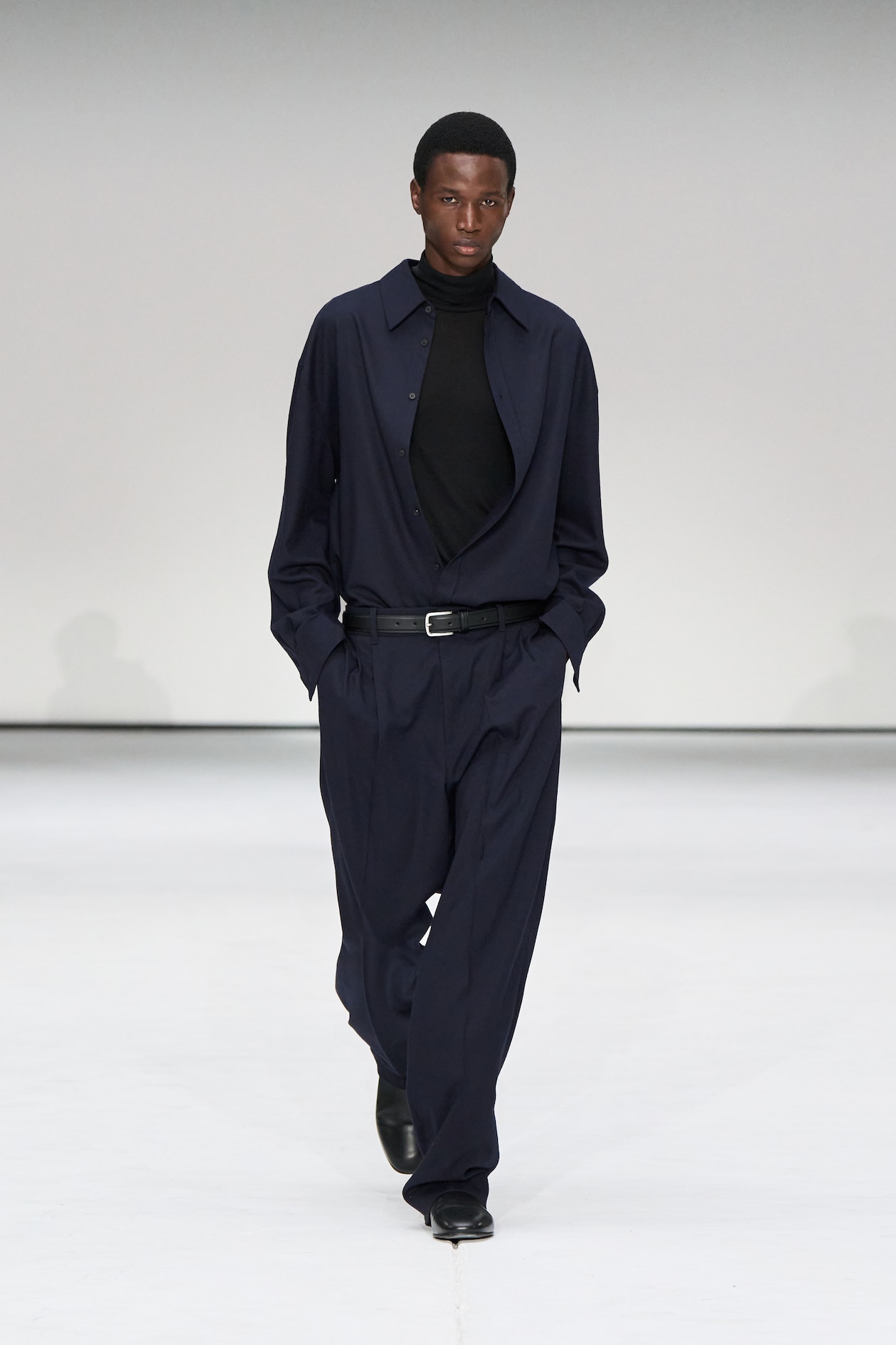 Model in a navy blue outfit with a black turtleneck walks confidently on a white runway, hands in pockets.