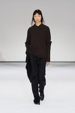 Placeholder for: Model in a dark brown sweater and black asymmetric skirt walks on a minimalist runway with a white background.
