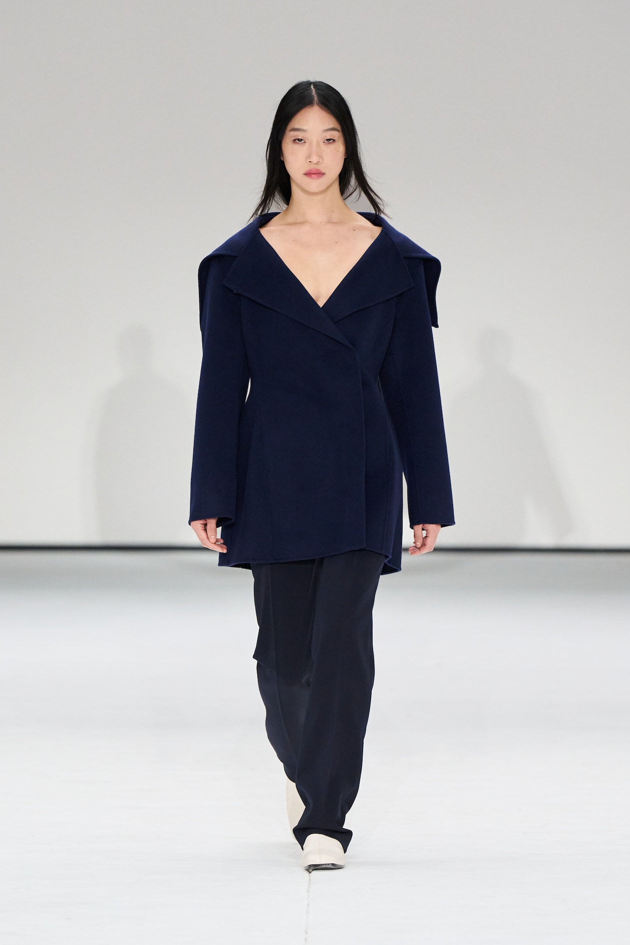 Model walking on a runway wearing a dark blue coat and pants, with white shoes, against a plain white background.