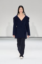 Placeholder for: Model walking on a runway wearing a dark blue coat and pants, with white shoes, against a plain white background.