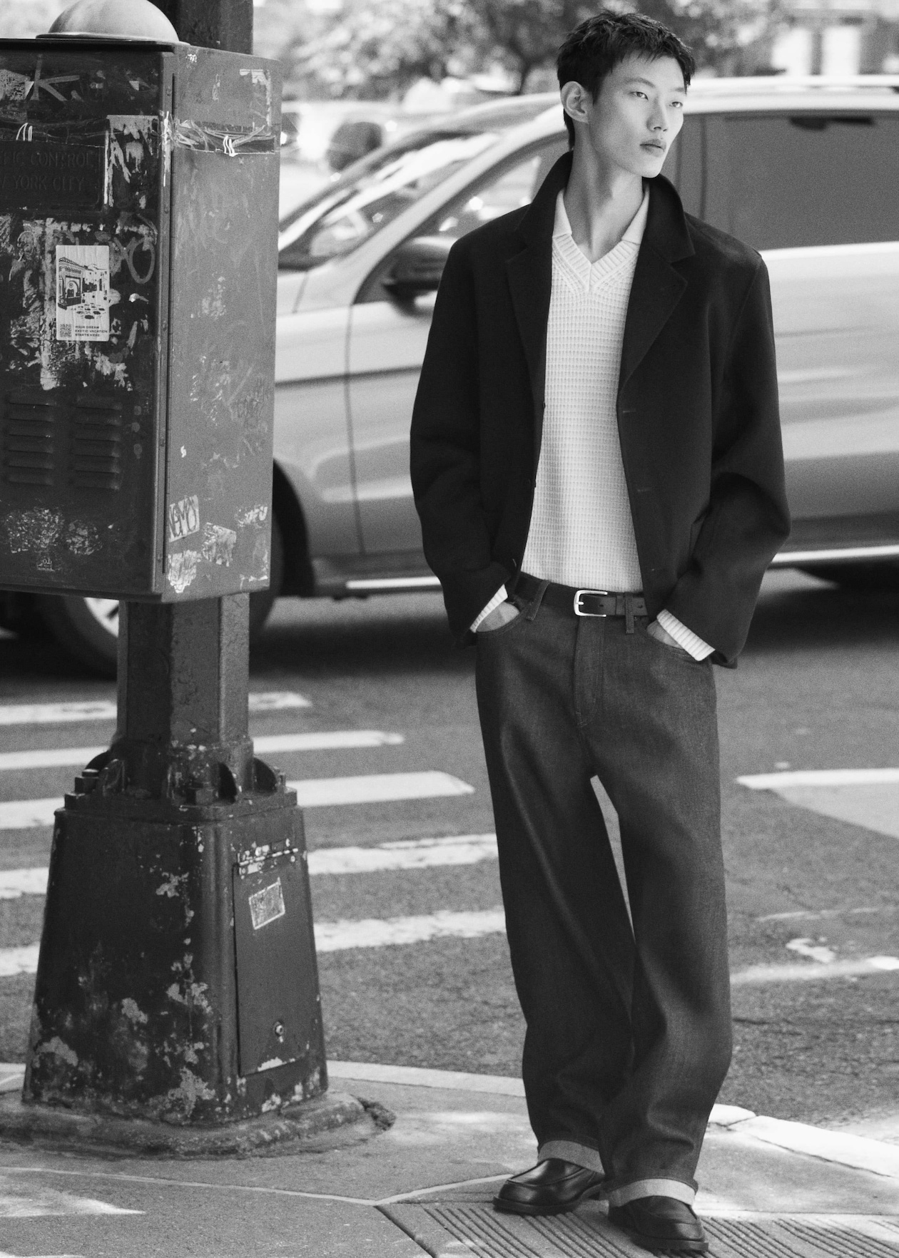 A person in a stylish outfit stands at a street corner in a black and white photo, with a blurred car in the background.