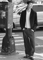 Placeholder for: A person in a stylish outfit stands at a street corner in a black and white photo, with a blurred car in the background.