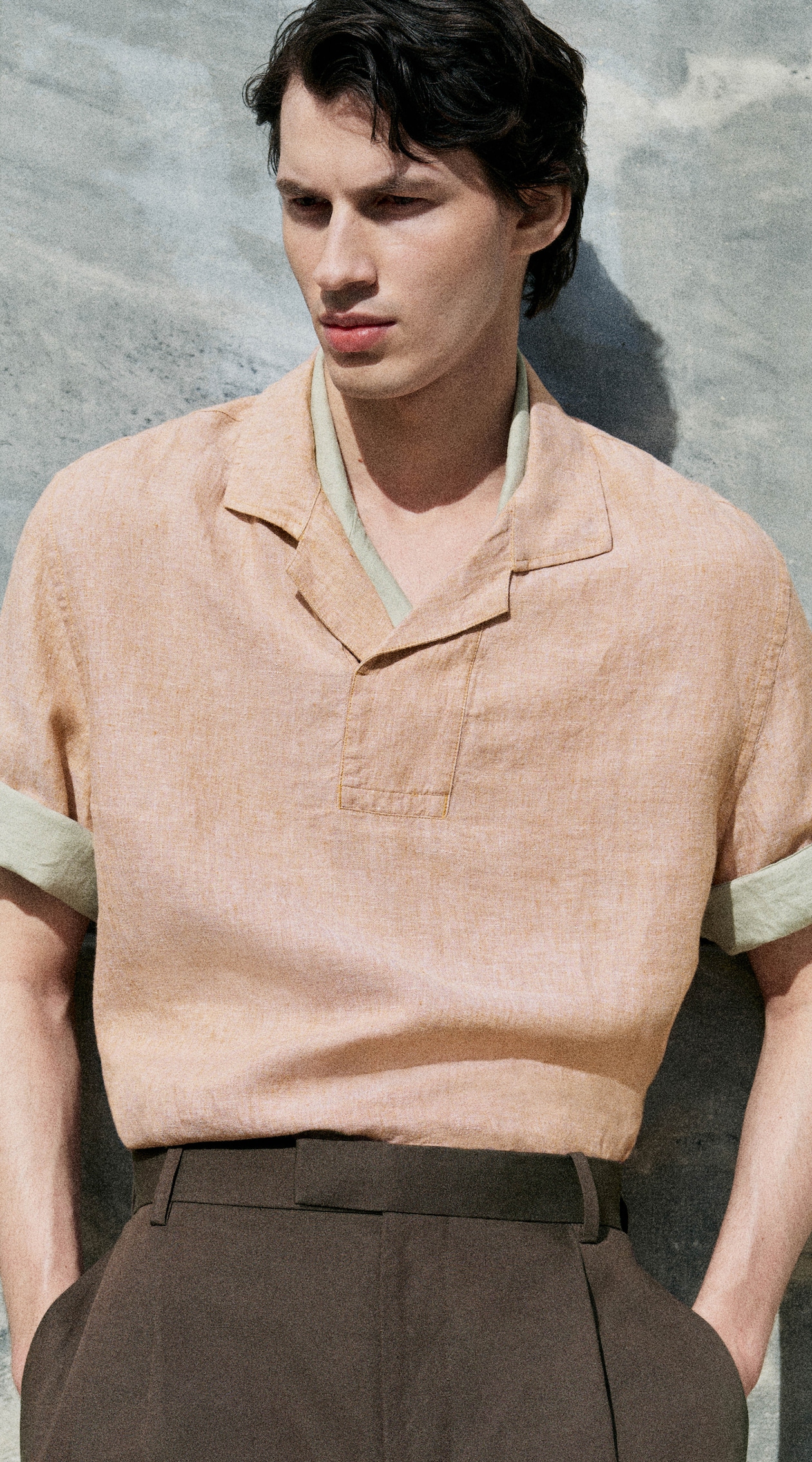 Man in a light orange shirt with rolled sleeves and dark trousers stands against a gray textured wall, hands in pockets, looking to the side.
