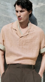 Placeholder for: Man in a light orange shirt with rolled sleeves and dark trousers stands against a gray textured wall, hands in pockets, looking to the side.
