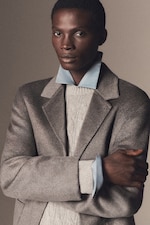 Placeholder for: A person wearing a light gray textured coat over a light-colored cable knit sweater with a blue shirt collar peeking out, posing with crossed arms.