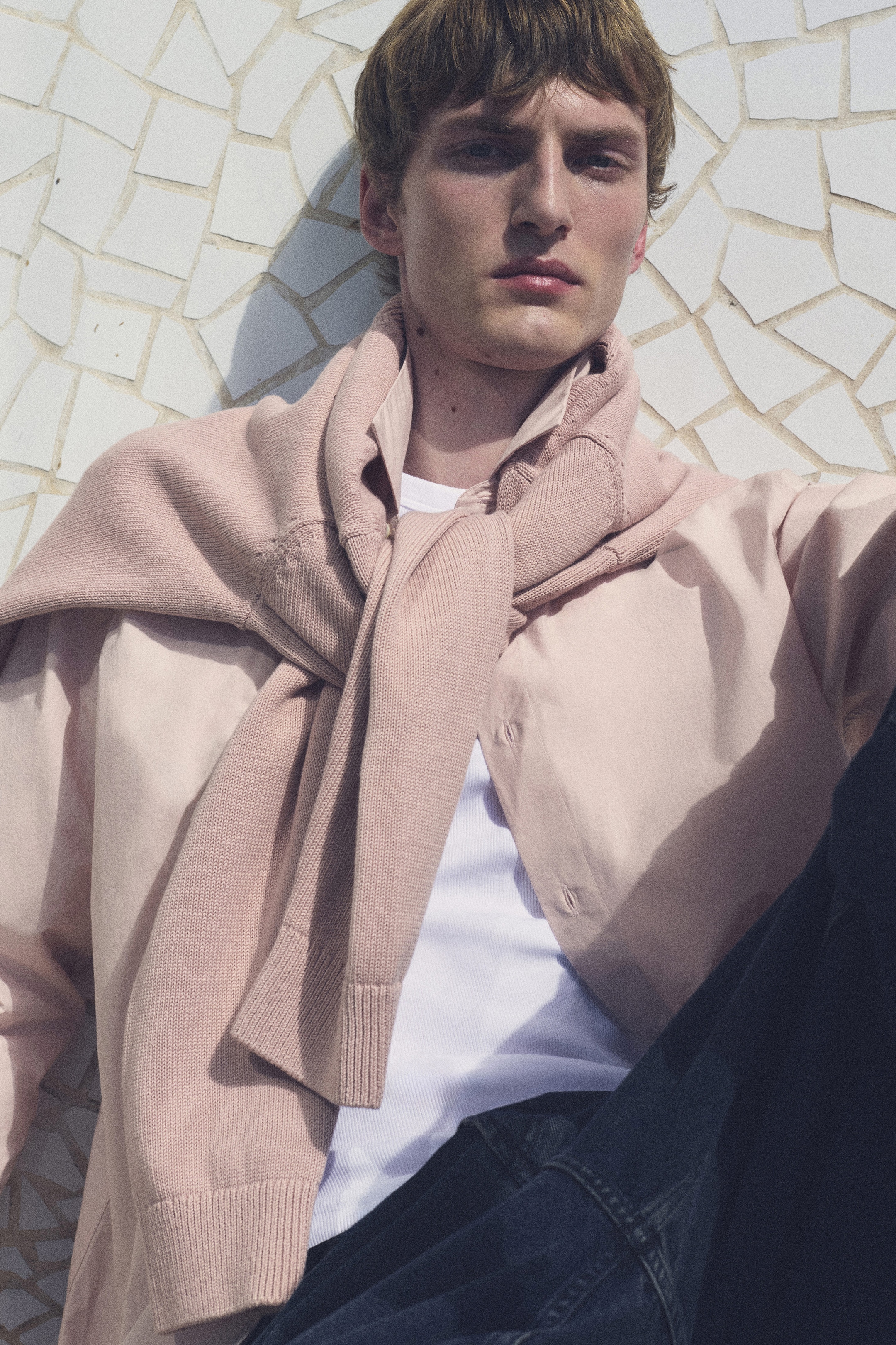 Young man with a pensive expression, wearing a light pink sweater over his shoulders and a white shirt, posed against a tiled wall.