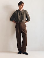 Placeholder for: Man in a grey cardigan and high-waisted brown trousers, standing against a plain white background, arms crossed and looking to the side.