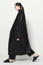 Placeholder for: Person in a long black coat and black pants, walking sideways, with hair in a sleek ponytail, wearing black slip-on shoes.