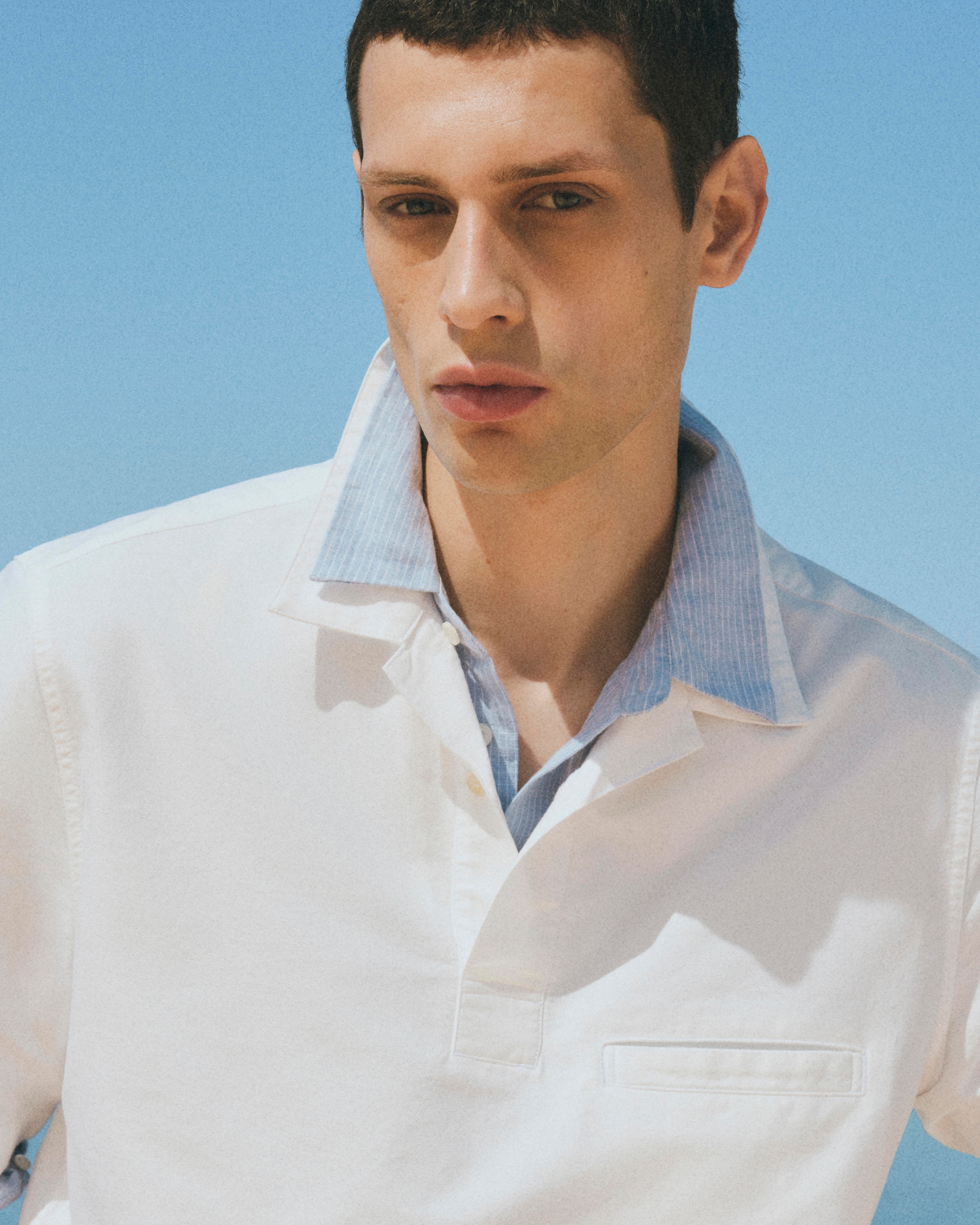 Man in a white shirt with a blue collar stands against a clear blue sky, looking at the camera with a neutral expression.