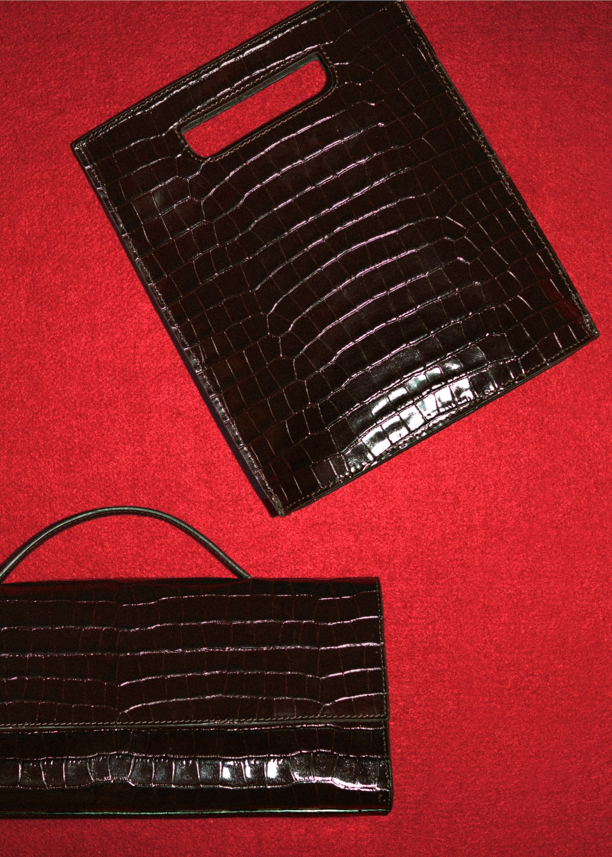 Two dark brown crocodile-patterned leather bags on a red surface; one with a handle slot, the other with a top handle.