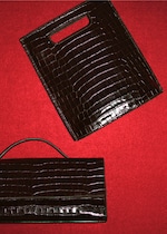 Placeholder for: Two dark brown crocodile-patterned leather bags on a red surface; one with a handle slot, the other with a top handle.