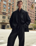 Placeholder for: A person wearing a black suit and standing confidently on a city street with tall buildings and a traffic light in the background.
