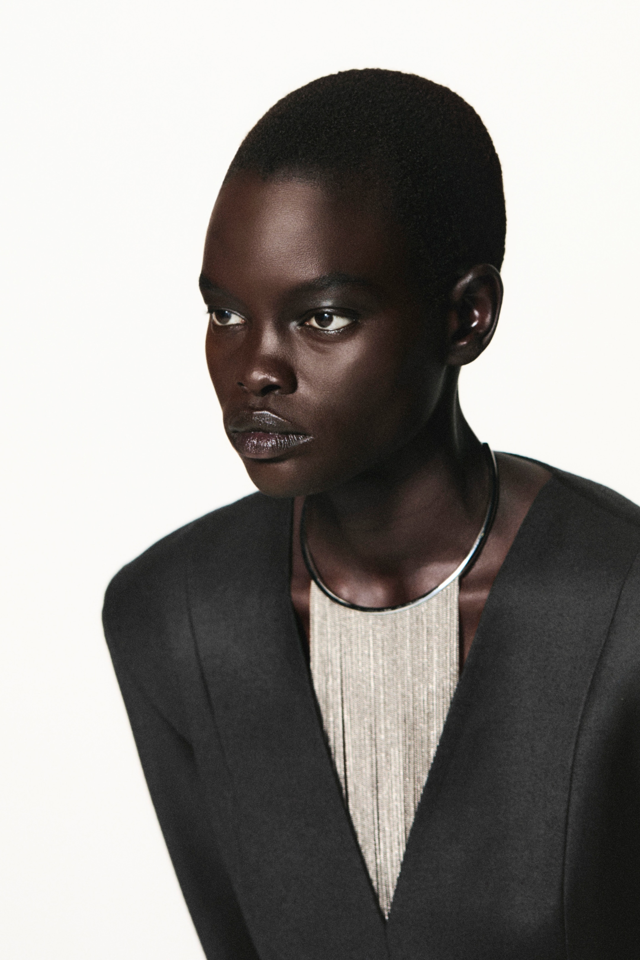 Person with short hair wearing a dark suit and necklace, gazing to the side against a plain background.