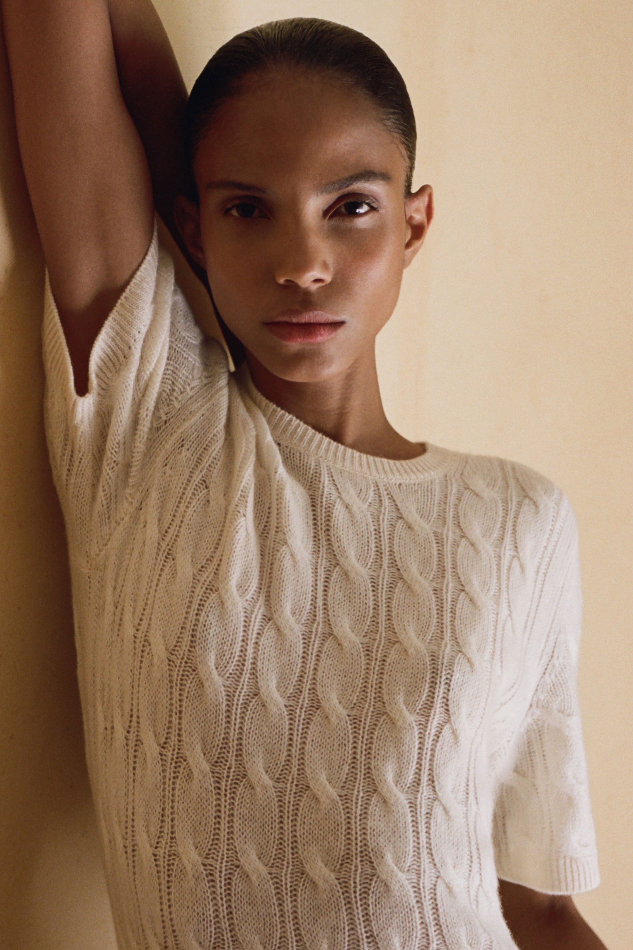 Person wearing a white cable knit sweater, leaning against a light-colored wall with one arm raised, looking directly at the camera.