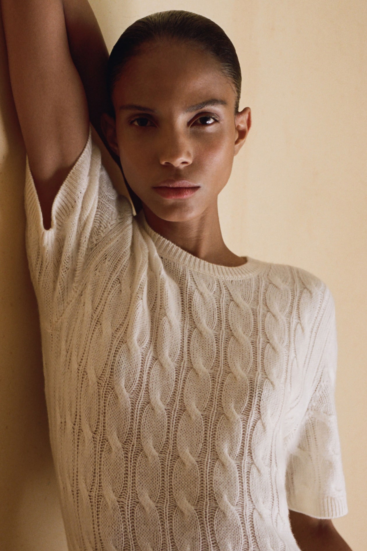 Person in a white cable-knit sweater standing against a beige wall, with one arm raised and looking directly at the camera.