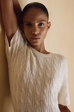 Placeholder for: Person in a white cable-knit sweater standing against a beige wall, with one arm raised and looking directly at the camera.