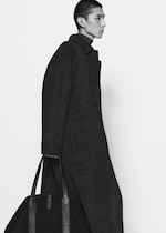 Placeholder for: Person in a long, dark plaid coat holding a large black bag, walking against a plain white background